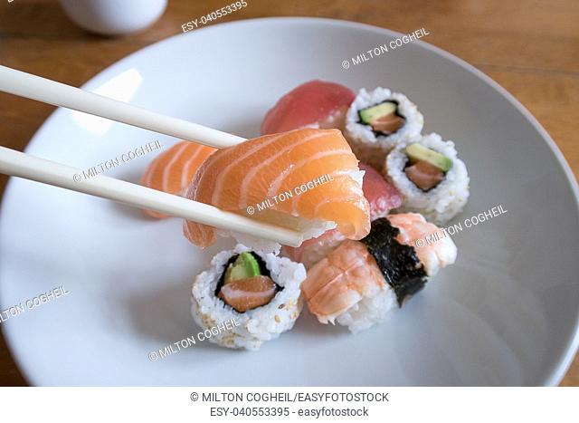 Fresh salmon nigiri sushi between chopsticks with uramaki, prawn nigiri and tuna nigiri in the background
