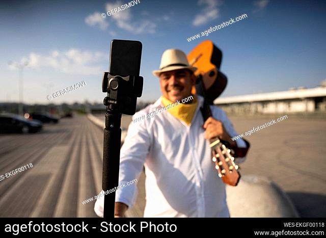 Smiling guitarist holding guitar taking selfie through mobile phone on tripod