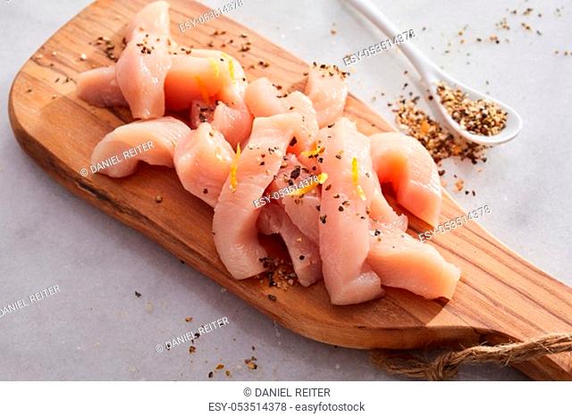Diced fresh raw chicken breast cut in strips for a goulash or stir fry seasoned with spice rub heaped on a wooden chopping board on white marble