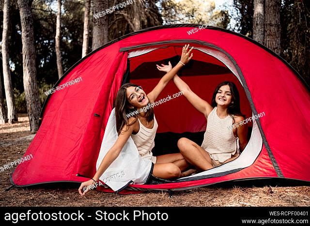 Cheerful sisters with hands raised sitting in tent at campsite during vacation