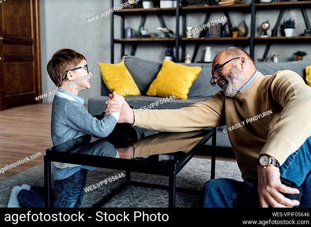 Happy senior man arm wrestling with grandson in living room at home