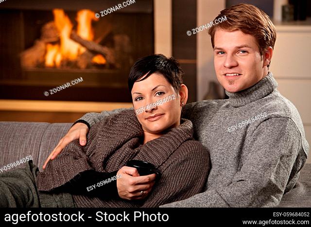 Young couple hugging on sofa in front of fireplace at home, looking at camera, smiling