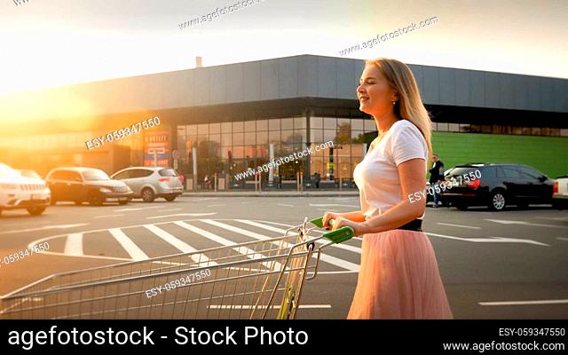 Beautiful smiling blonde girl pushing shopping cart while walking on car parking at shopping mall