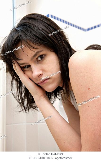 This picture shows a young caucasian woman with brown hair as she sits near her toilet feeling sick / ill in her bathroom