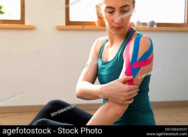 Young fit women checking her shoulder with elastic therapeutic kinetic tape. Performing exercise at home. Kinesiology physical therapy