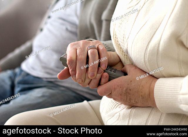 Elderly woman holding television remote with both of her hands. Aged grandma wearing white sweater holding tv remote with her husband next to her