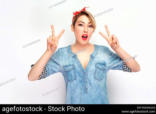 Portrait of funny beautiful young woman in casual blue denim shirt with makeup and red headband standing with victory or peace sign and looking at camera