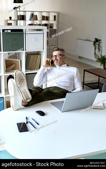Smiling businessman with feet up talking on mobile phone in office