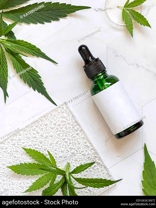 Dropper bottle with blank label near green cannabis sativa leaves on a marble table top view. Cosmetic Mockup, Copy space
