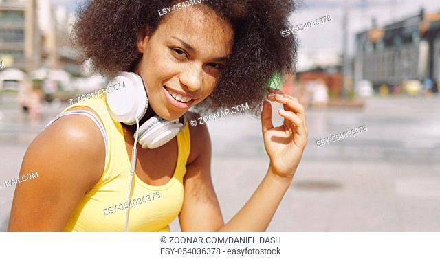 Portrait of young woman in yellow tank top wearing white headphones on neck and looking at camera on urban background