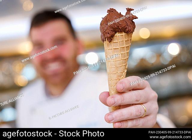 PRODUCTION - 23 March 2023, Bavaria, Schwabach: Ice cream maker Luca de Rocco holds a waffle cone with a scoop of vegan chocolate ice cream in his ice cream...
