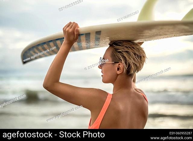 Delightful blonde girl with short hairstyle stands backsides on the beach on the background of the sea and cloudy sky. She wears orange swimsuit with sunglasses...