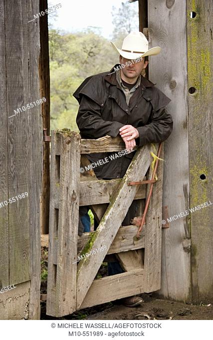 Cowboy up against gate and old rugged barn in the country on a ranch looking downward in a relaxed style