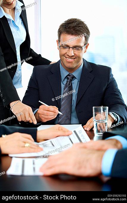 Smiling executive signing contract in office, assistant pointing at paper