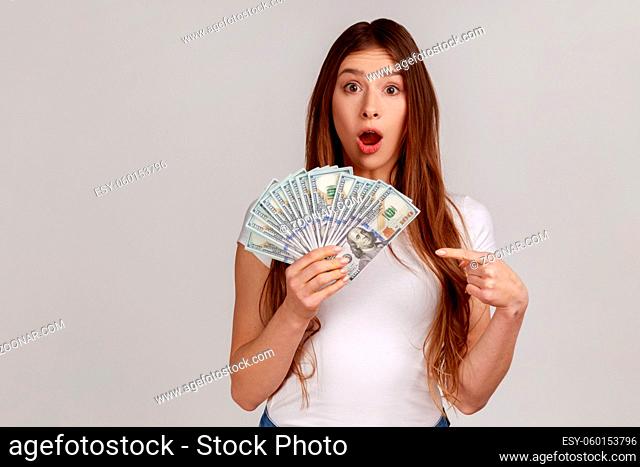 Portrait of woman holding pointing at fan of dollar banknotes, looking at camera with astonishment, wearing white casual style T-shirt