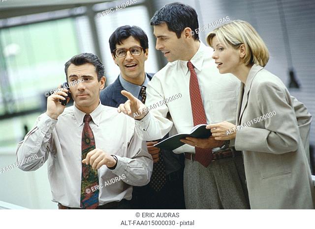 Four business colleagues standing in a group, one using phone, others listening