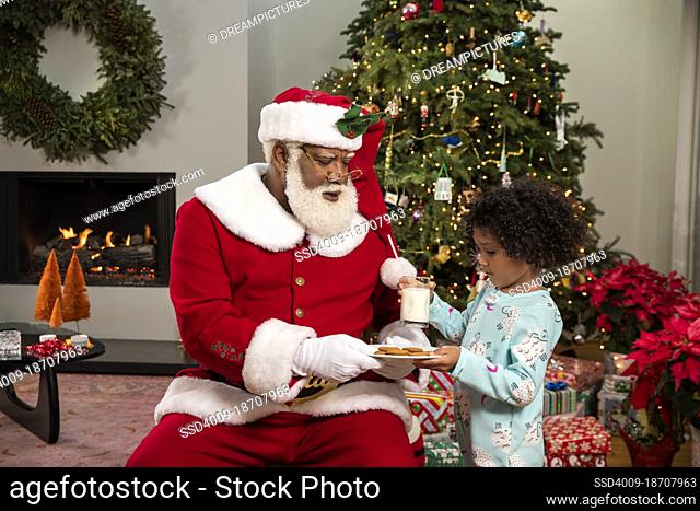 Cute young girl handing Santa Claus milk and cookies in front of Christmas tree
