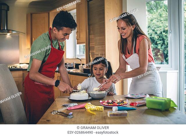 Mother decorating a cake with her son and daughter in the kitchen of their home. They are covering the cake with icing and have sweets to decorate