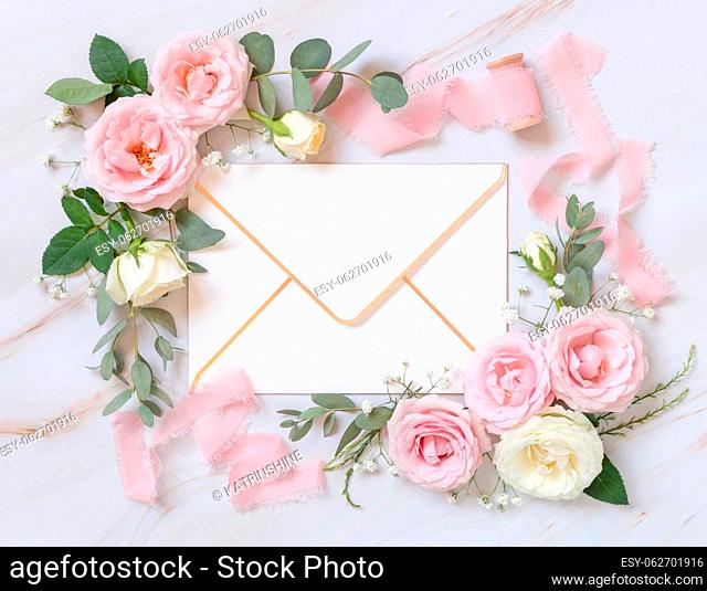 Paper envelope between light pink roses and silk ribbons on marble top view, wedding mockup. Romantic scene with blank envelope and pastel flowers flat lay
