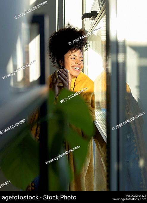 Mujer sonriente con taza de café mirando por la ventana en casa