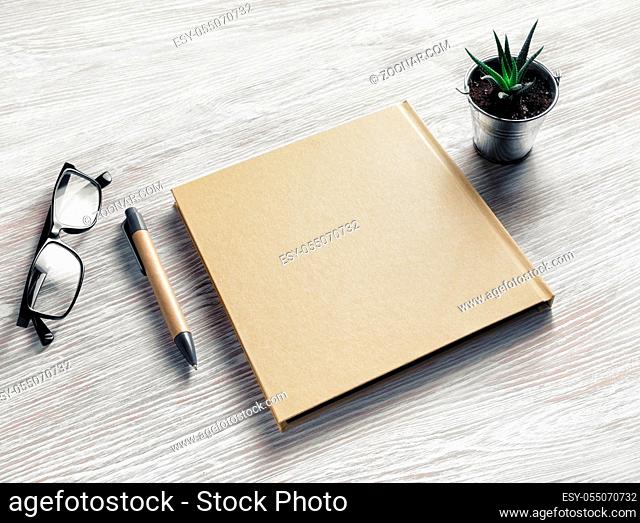 Photo of closed blank sketchbook, glasses, pen and plant on light wood table background. Blank stationery template