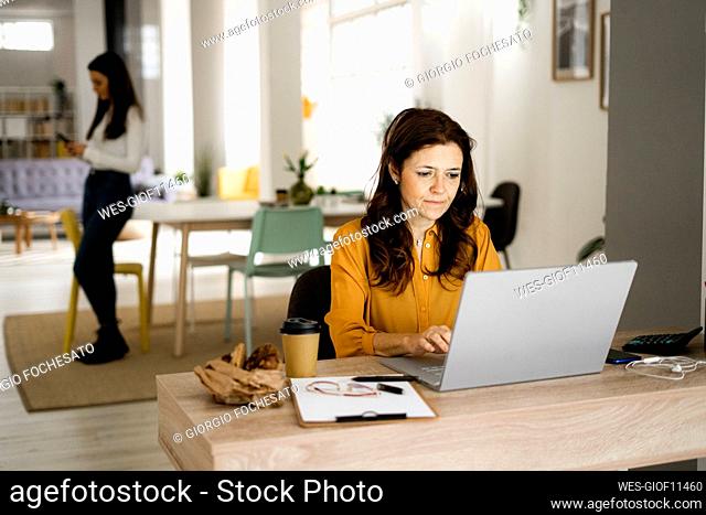 Mature businesswoman working on laptop while daughter using mobile phone at home office