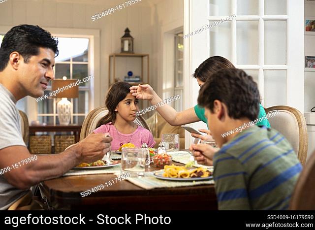 Hispanic family around the dinner table with the daughter feeling ill, mom checking her temperature and checking cell phone to connect with doctor