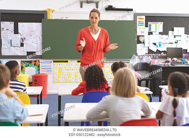 Front view of a teacher teaching to elementary students in classroom at school with greenboard in background