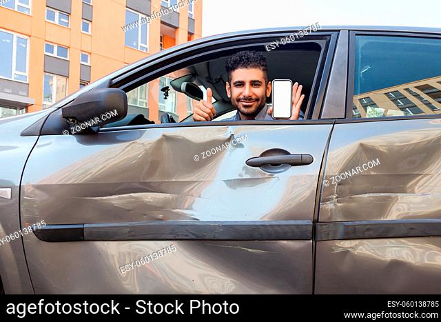 Portrait of bearded man in crashed car looking at camera and smiling, showing thumb up and smart phone with blank screen