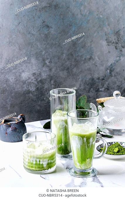 Matcha green tea iced latte or cocktail in three different glasses with ice cubes, matcha powder and jug of milk on white marble table