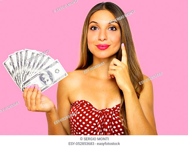 Young thinking pretty woman in strapless dress holding a bunch of money banknotes, looks at camera as she has an idea how to spend it