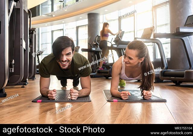 Cheerful young couple exercising forearm plank side by side on mats during workout in the interior of a modern fitness club
