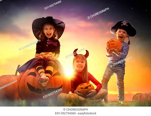 Happy brother and two sisters on Halloween. Funny kids in carnival costumes outdoors. Cheerful children and pumpkins on sunset background