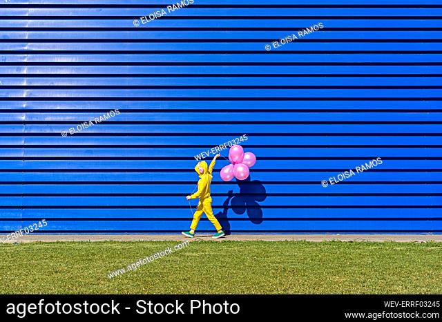 Little girl with headphones wearing yellow tracksuit walking in front of blue background with pink balloons