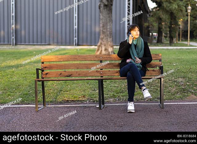 Latin woman sitting on a bench in a park talking on a cell phone