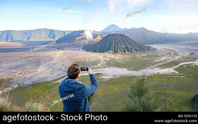Young man takes photo with iPhone, volcanic landscape, view in Tengger Caldera, smoking volcano Gunung Bromo, in front Mt. Batok, in the back Mt