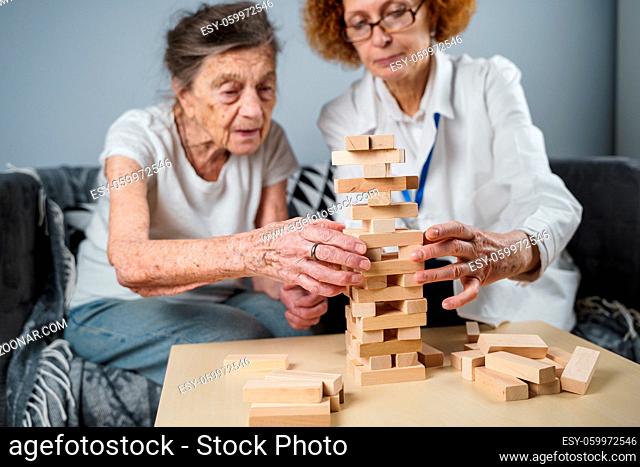 Jenga game. Theme is dementia, aging and games for old people. Caucasian senior woman builds tower of wooden blocks with the help of a doctor as part of a...