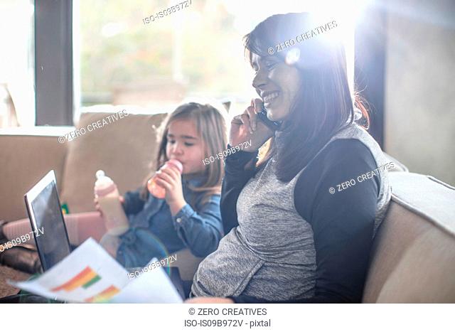 Girl with baby bottle while mother working on sofa making smartphone call