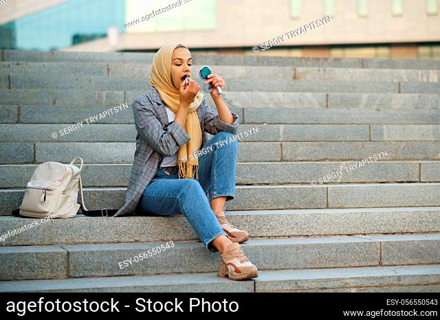 Arab female student sitting on the stairs in downtown. Muslim woman applies makeup at the entrance to the business center. Religion and education