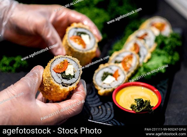 PRODUCTION - 07 November 2023, Lower Saxony, Oldenburg: A chef holds a sushi roll with kale, tomatoes and feta in his hands in the kitchen of the Moto Kitchen...