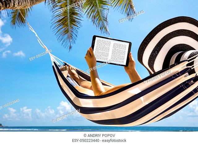 Woman In Hat Relaxing On Hammock Reading The Book On Digital Tablet Against Blue Sky