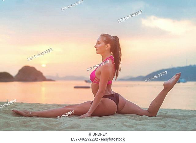 Photo in profile of slim fitness model with brown hair wearing bikini doing leg split exercise on beach at sunrise against sea, sky and mountains