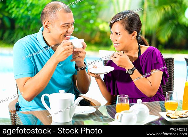 Asian couple having coffee on the porch in front of their home, in the background a tropical garden