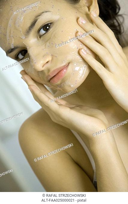 Young woman applying facial mask, looking at camera, cropped view
