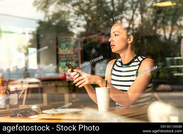 Thoughtful caucasian woman holding mobile phone while looking through the coffee shop window during coffee break. Street reflections in the window glass