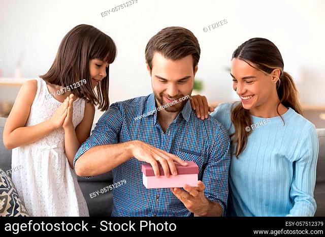 Excited young father sit on couch with wife and little daughter open birthday surprise, happy mom and preschooler girl kid make present give gift box to smiling...
