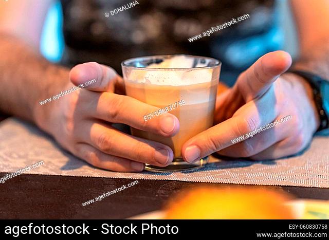 Selective focus on the hands of a man holding a glass of cappuccino, spreading his index fingers
