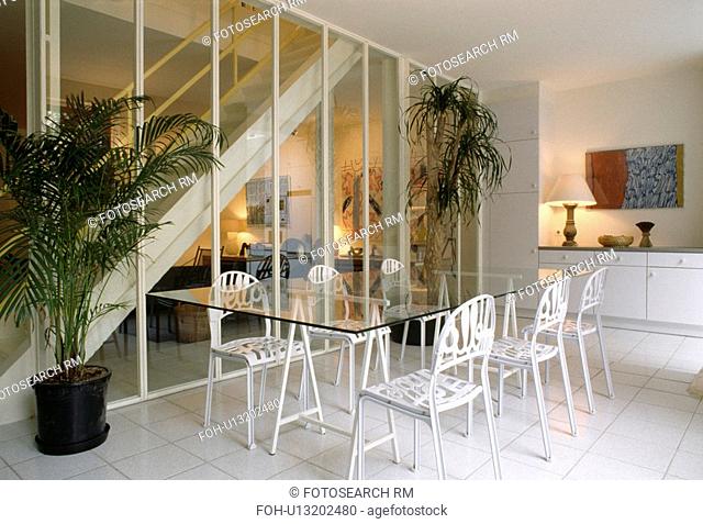 White metal chairs and glass trestle table in front of clear glass wall in hall dining room with white tiled floor