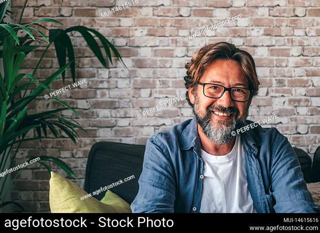 Portrait of handsome happy mature man looking at camera and smiling in the living room of modern apartment. Confident guy with with eyeglasses sitting...