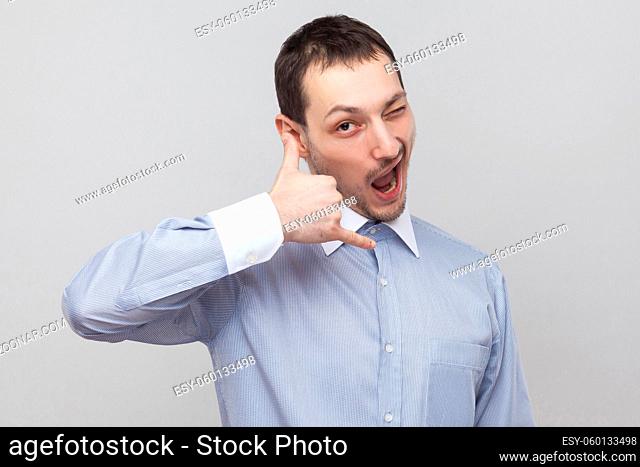Portrait of funny handsome bristle businessman in classic light blue shirt standing with call gesture and looking at camera winking